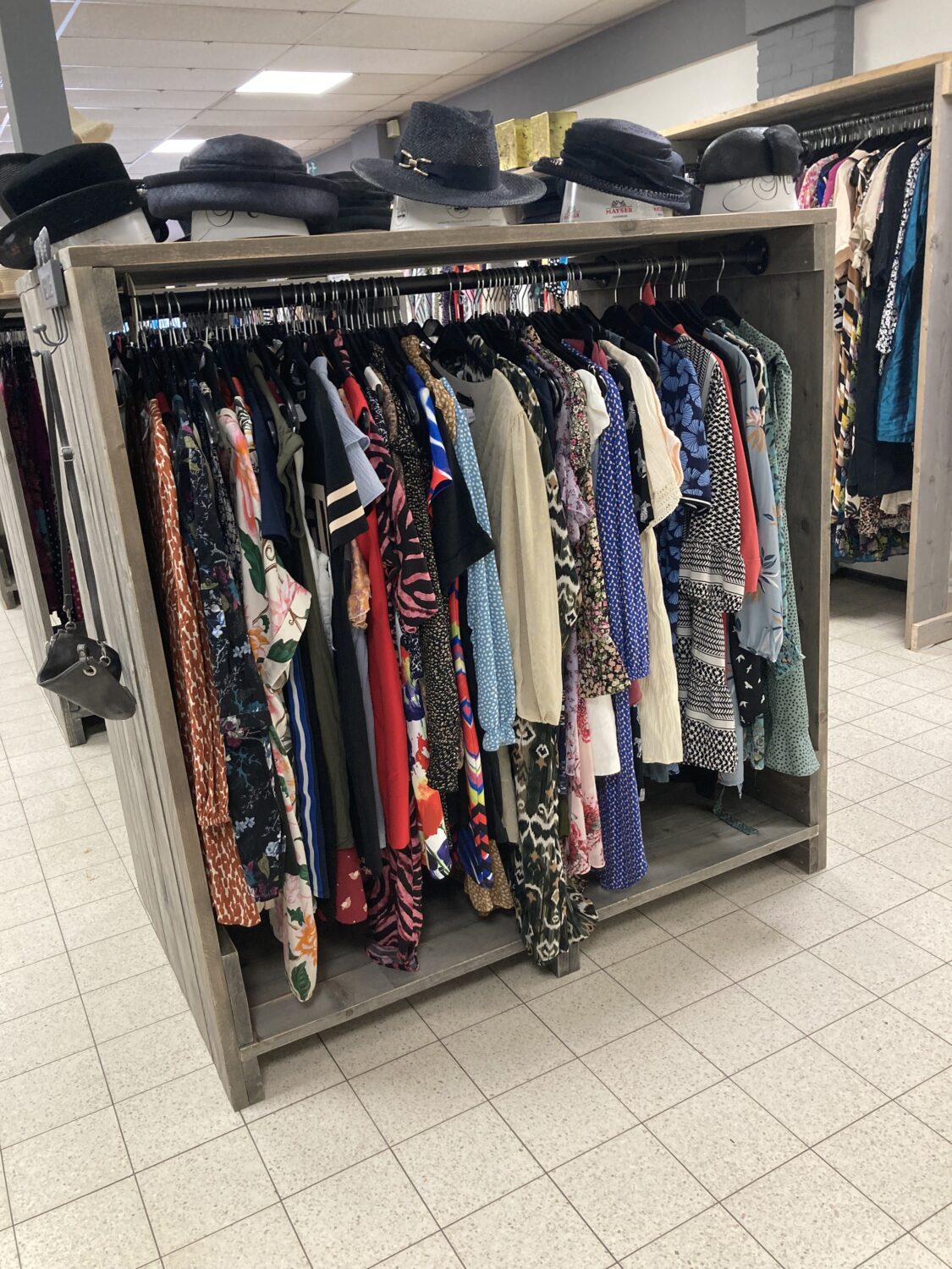 Een kledingrek gevuld met verschillende tweedehands kledingstukken en hoeden in een winkel of kringloopwinkel.