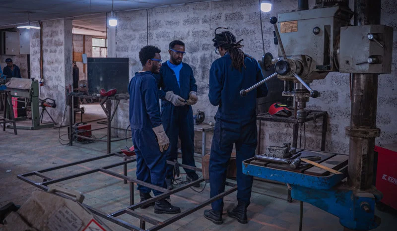 Drie technici in werkplaats bespreken een project, omringd door industriële machines en gereedschap.