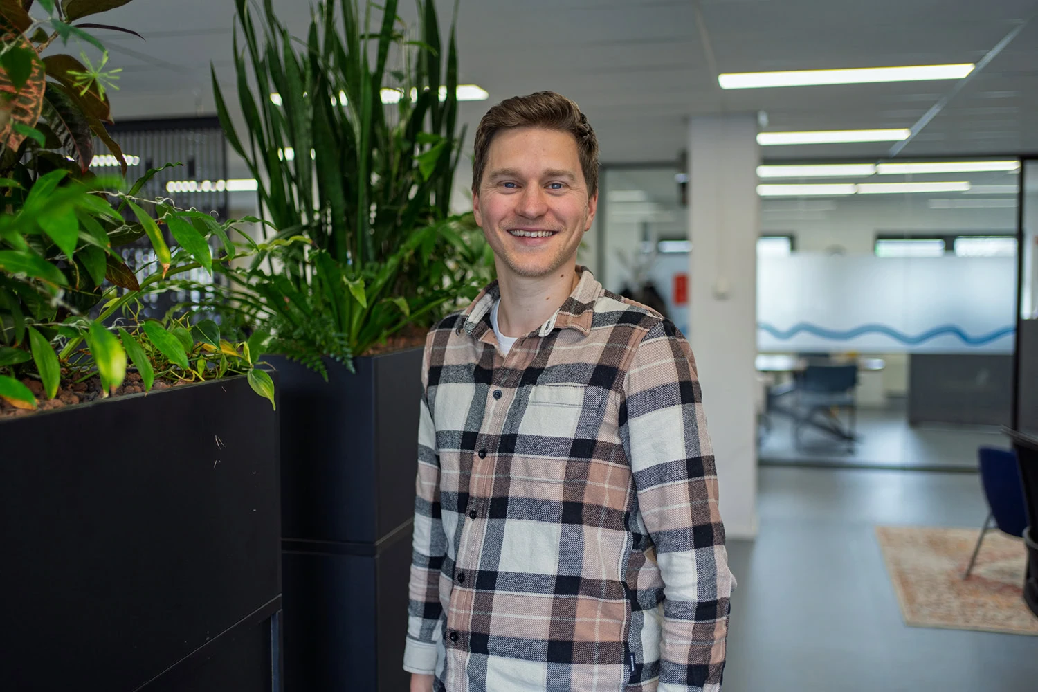 Een man glimlachend in een modern kantoor, omringd door groene planten en werkplekken, geschikt voor zakelijke en professionele doeleinden.