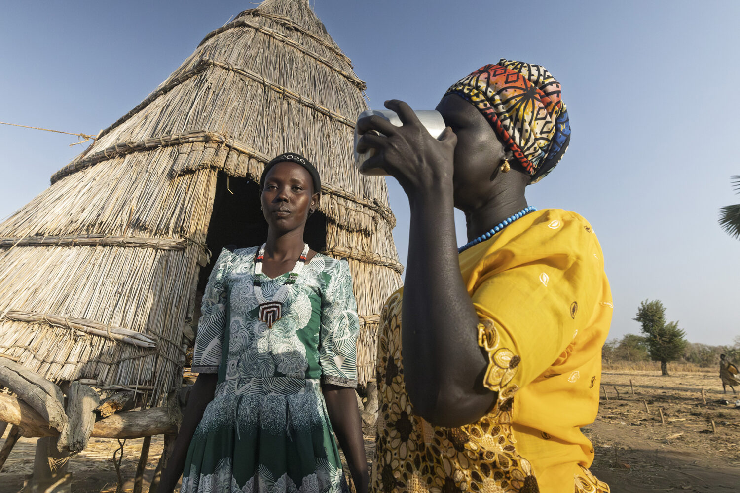 Nyanut en haar kinderen in een dorpsomgeving, met een traditionele hut op de achtergrond, terwijl ze water drinken uit een beker, illustrerend de strijd tegen schoon drinkwater.
