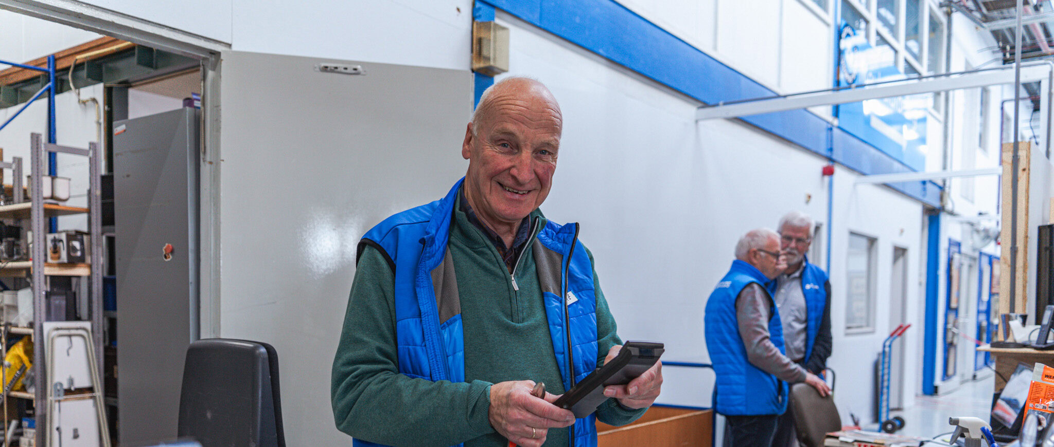 Een vrijwilliger controleert een apparaat in de winkel van Dorcas, omringd door andere vrijwilligers die helpen met de organisatie en verkoop van goederen.