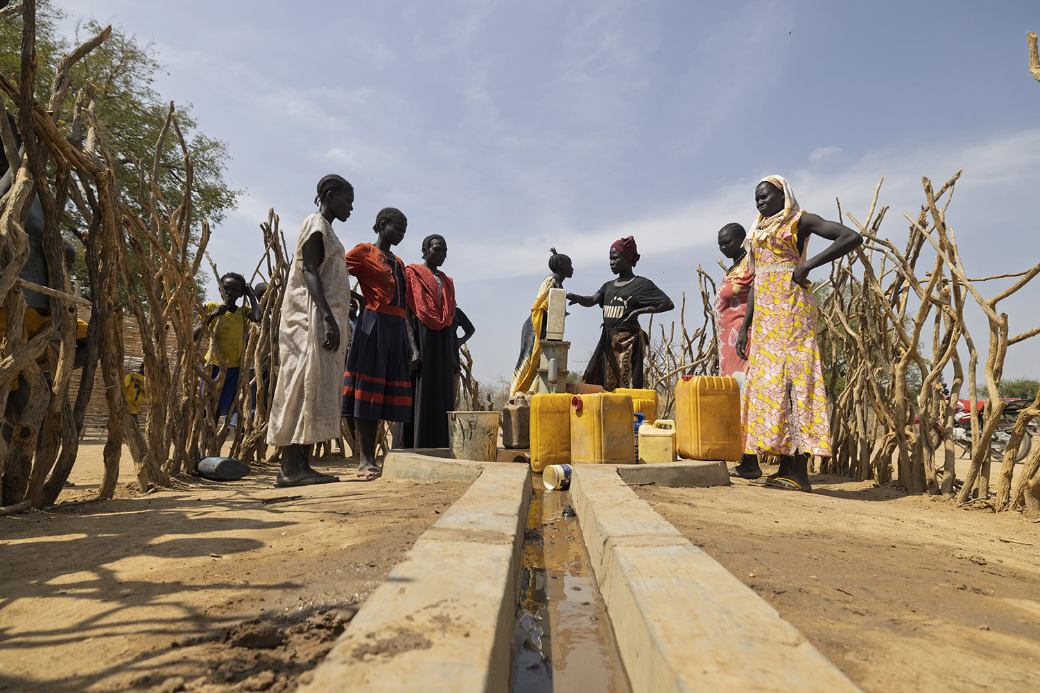 Gezinnen in Afrika gebruiken een waterpunt met een waterleiding, omgeven door houten palen en een groep mensen die water halen, onder een heldere blauwe hemel.
