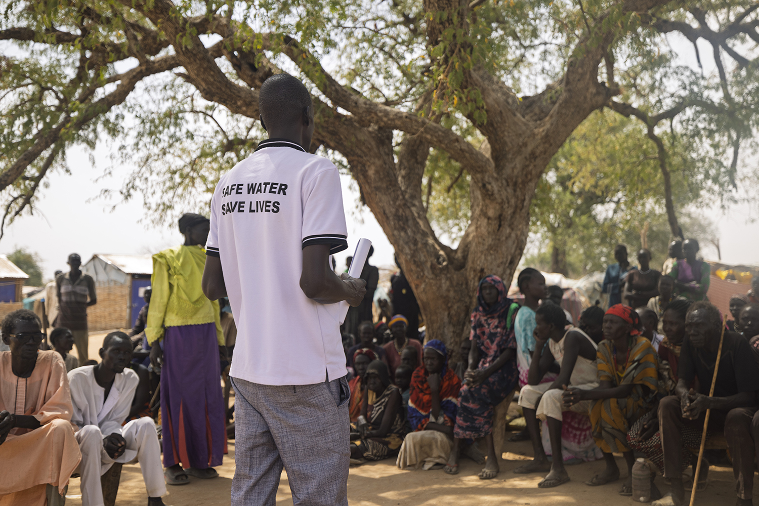 Een man geeft een presentatie over waterprojecten aan een groep mensen onder een grote boom in een dorre omgeving.
