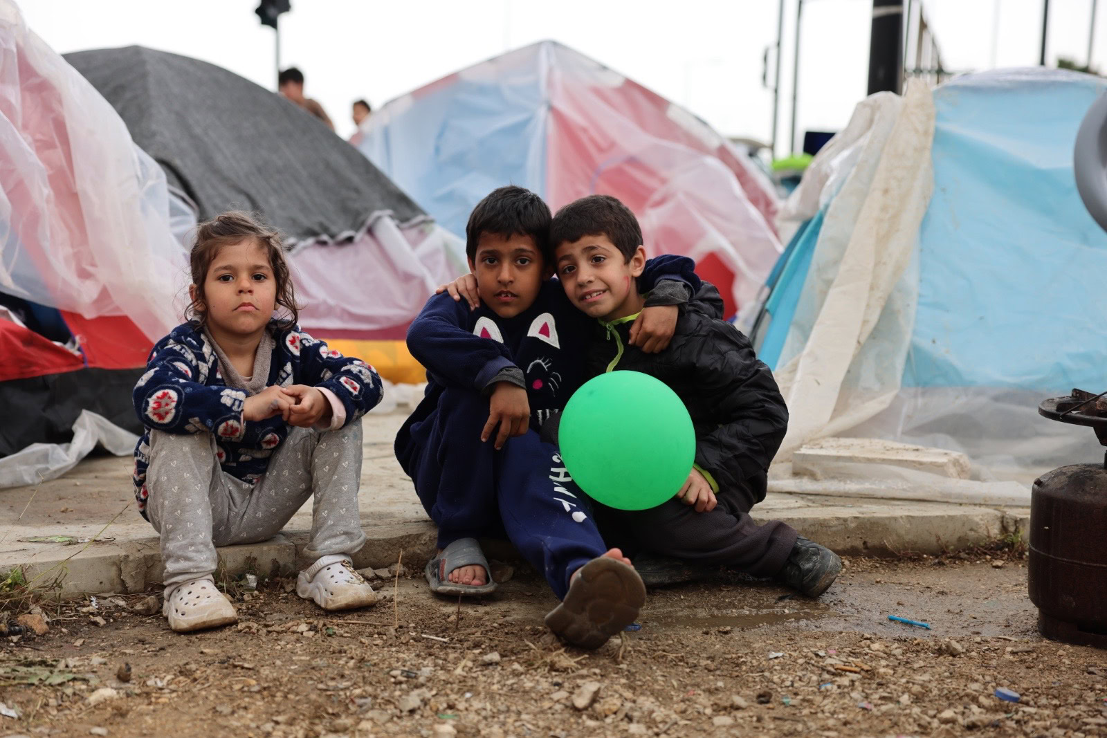 Drie kinderen zitten op de grond tussen tenten in een vluchtelingenkamp, met één kind dat een groene ballon vasthoudt, en zien er vermoeid maar hoopvol uit.
