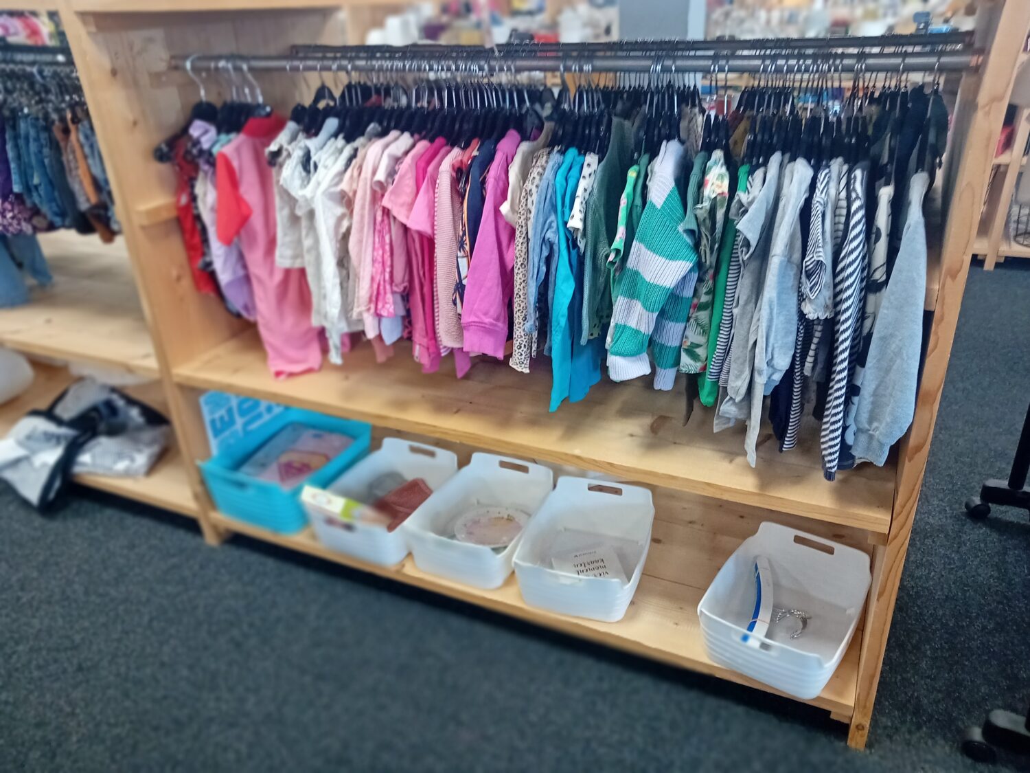 Een kledingrek met diverse kinderkleding in verschillende kleuren en patronen, aangevuld met opbergdozen onder het rek voor extra organisatie.