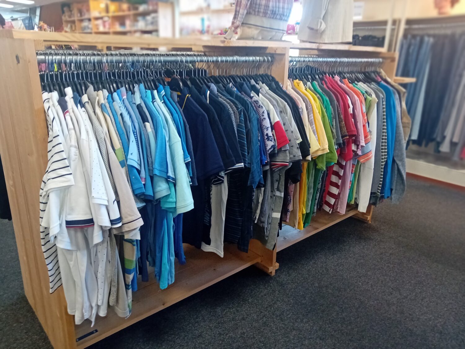 Kinderkleding op een houten rek in een winkel, met diverse shirts en t-shirts in verschillende kleuren en patronen, geschikt voor kinderen.