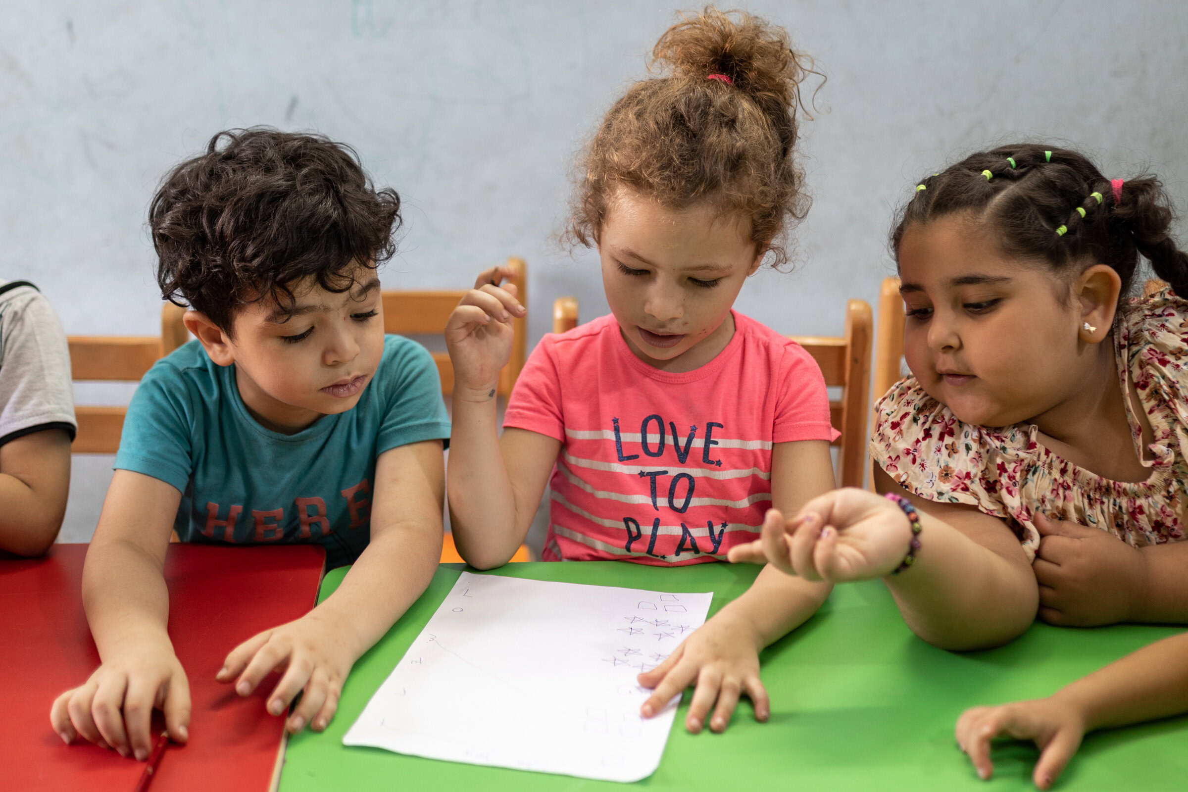 Drie jonge kinderen werken samen aan een schoolproject aan een groene tafel, wat samenwerking en leren bevordert.