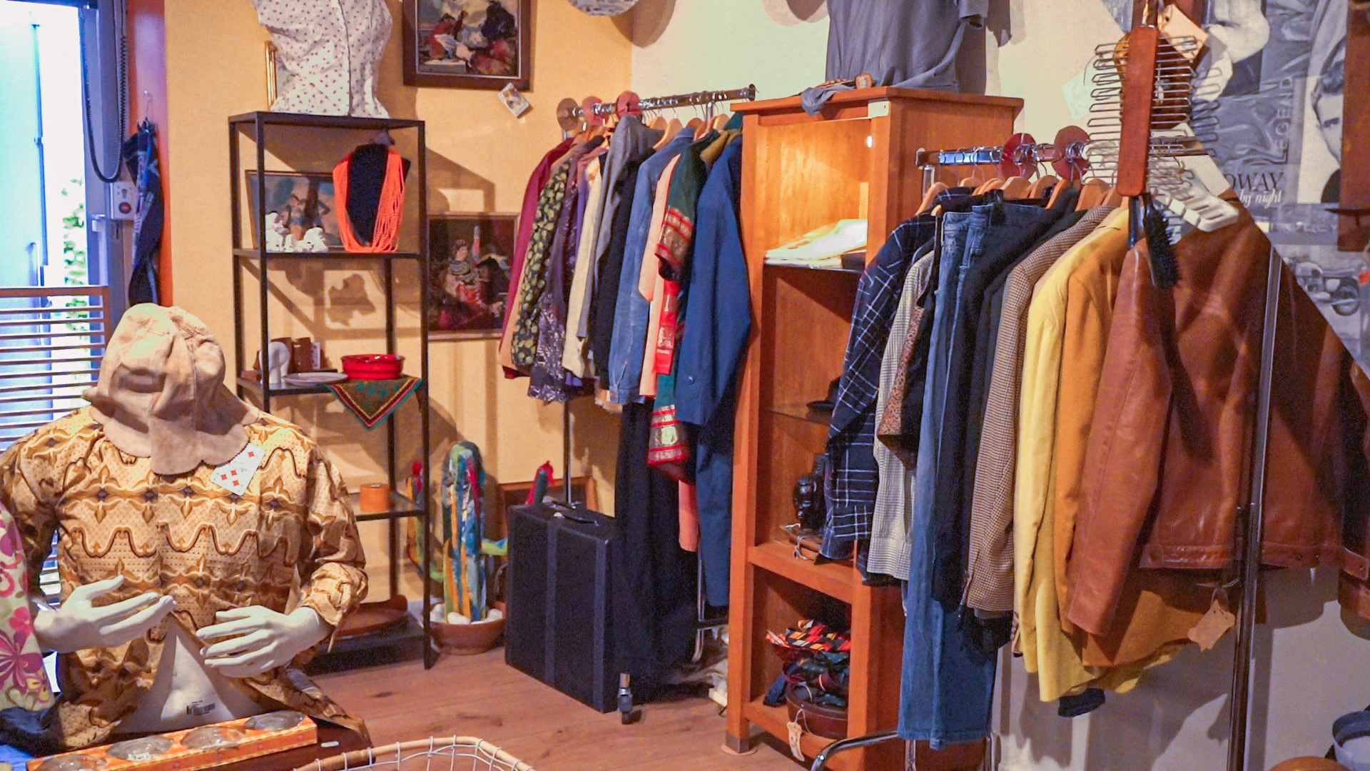 Vintage kledingwinkel met rekken vol jassen, jeans en blouses. Op de voorgrond staat een paspop met een beige patroonjurk en een hoed. In de winkel hangen verschillende kledingstukken en accessoires in een warme, sfeervolle inrichting.