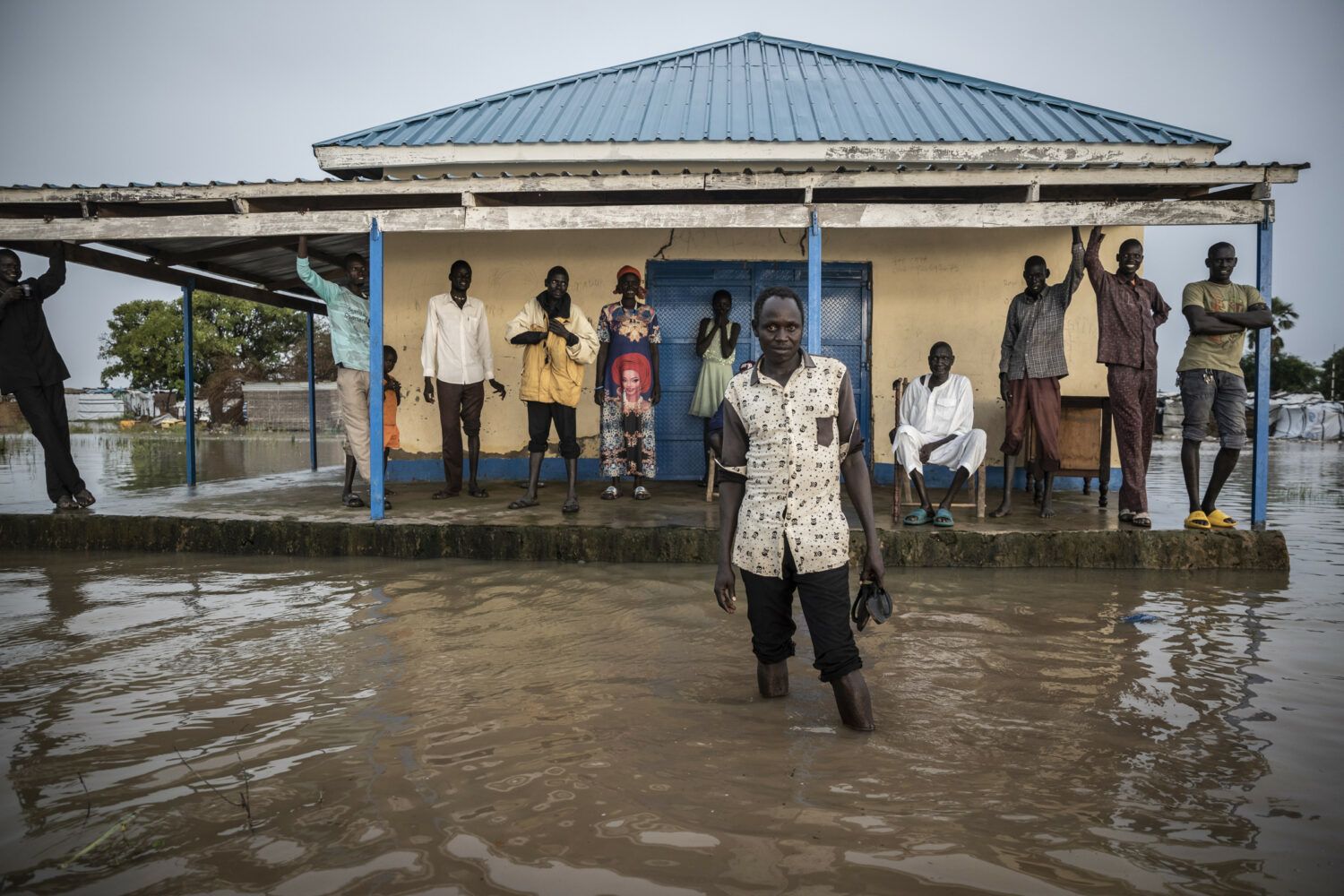 Mensen staan op een verhoogde veranda in een door water omringd dorp, symboliseert hulp en hoop in overstromingsgebieden in landen waar we hoop brengen.