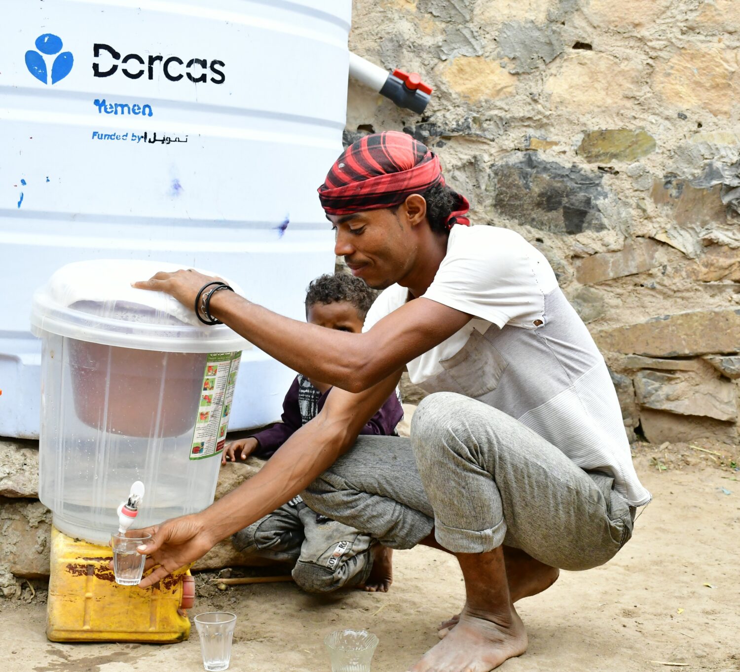 Man vult water uit een waterpunt met een jonge jongen op de achtergrond, in Jemen, voor een project van Dorcas.