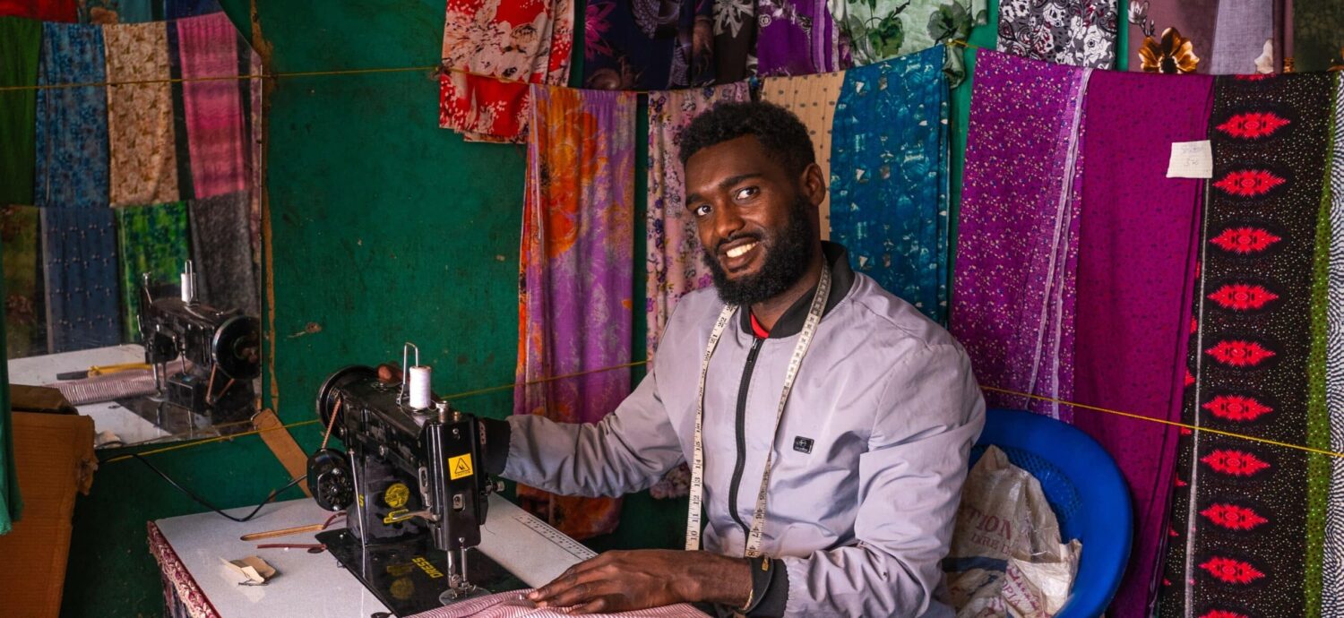 Man met een glimlach achter een naaimachine, omringd door kleurrijke stoffen en textielproducten, in een creatieve werkruimte voor textielproductie.