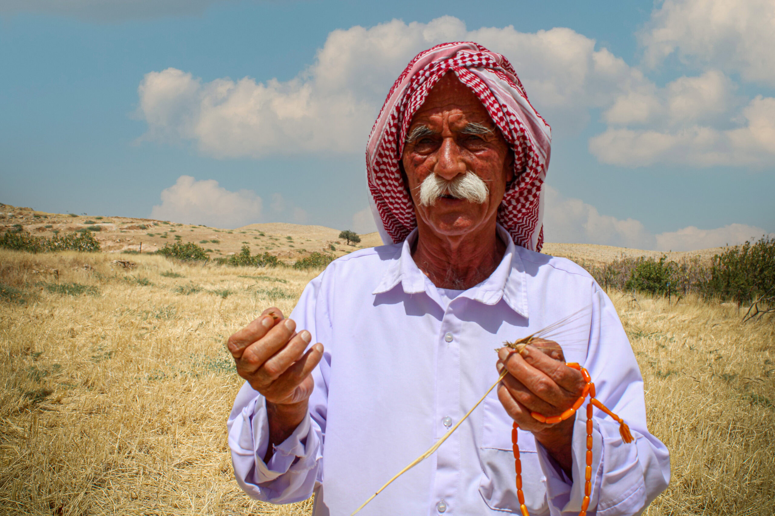 Irakse man met traditionele kleding en hoofddoek in een uitgestrekt veld, mogelijk tijdens een culturele of landelijke activiteit, onder een heldere blauwe hemel.