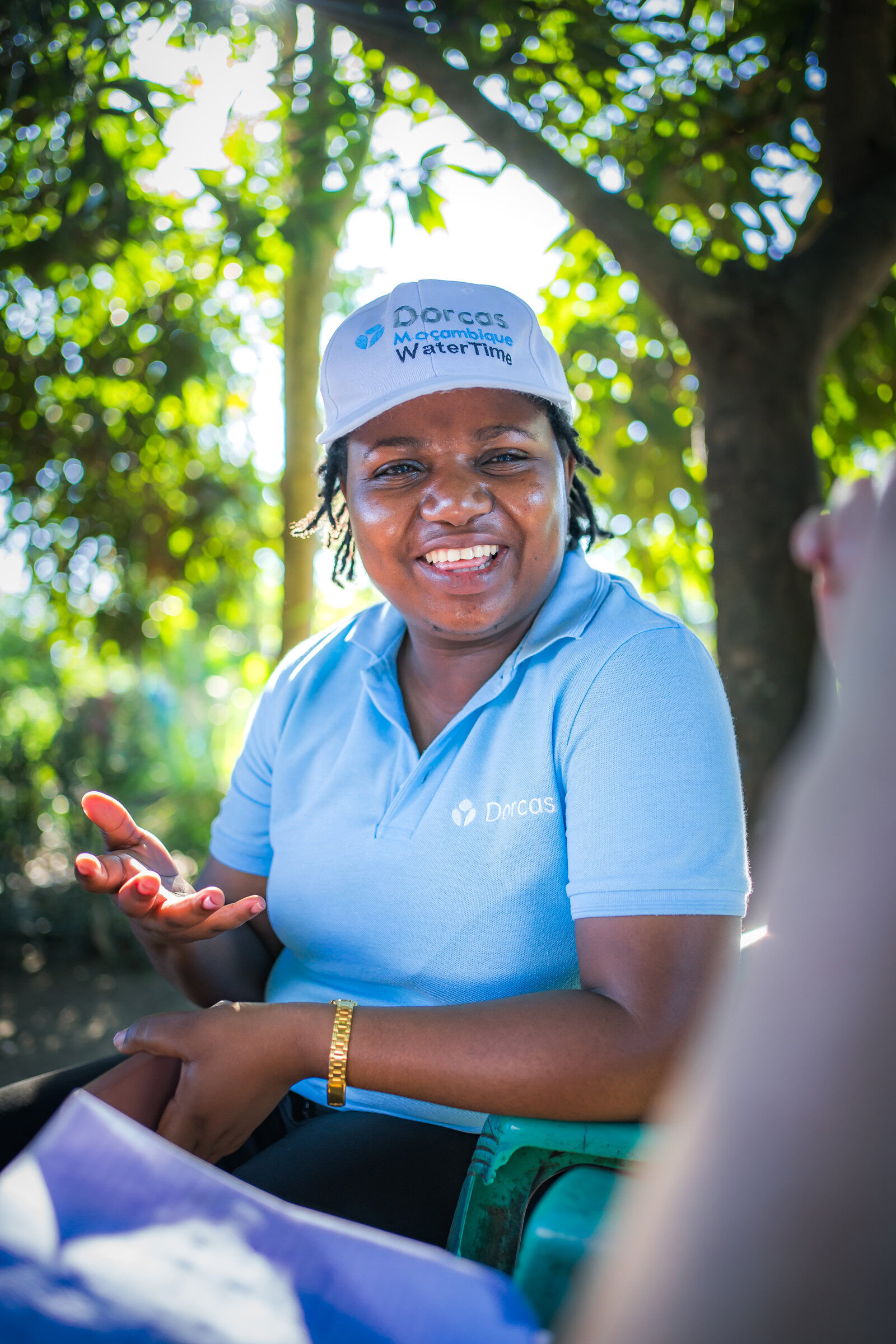 Vrolijke Dorcas-medewerker in blauwe uniform en pet met CHS Keurmerk, lachend en in gesprek, buiten onder groene bomen, symbool voor betrouwbare waterprojecten en kwaliteitszorg.
