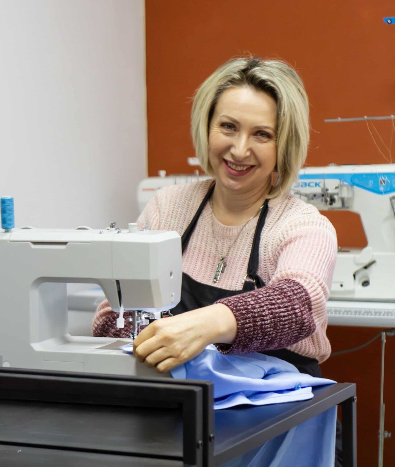 Vrouw met blond haar en een glimlach gebruikt een naaimachine in een naaiatelier, omgeven door stoffen en naaiapparatuur, gericht op textielproductie en ambacht.