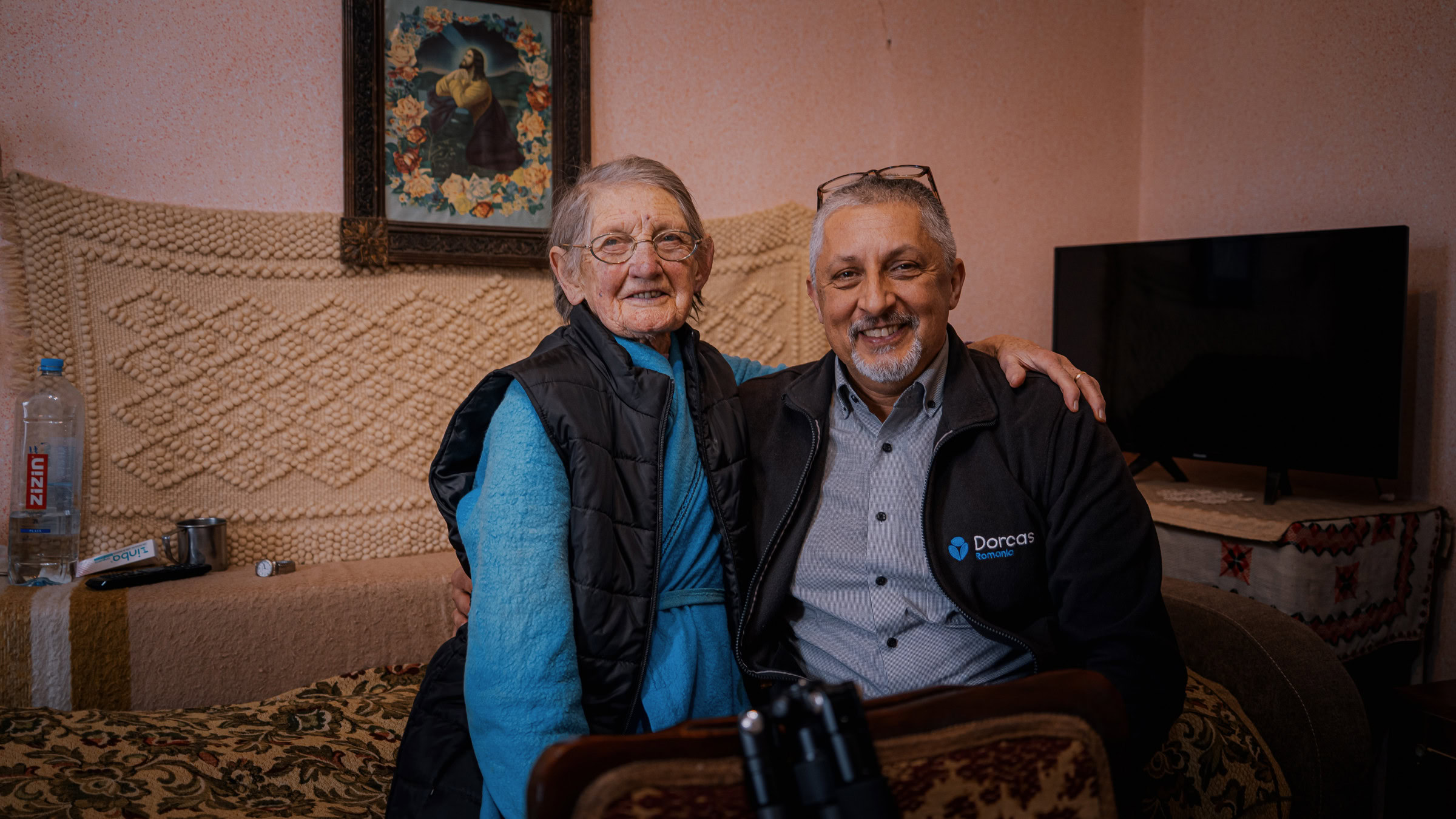 Vriendelijke oudere vrouw en man die samen lachen, zitten op een bank in een huis, met een Dorcas-jas zichtbaar, symboliseert betrokkenheid en zorg binnen de gemeenschap.