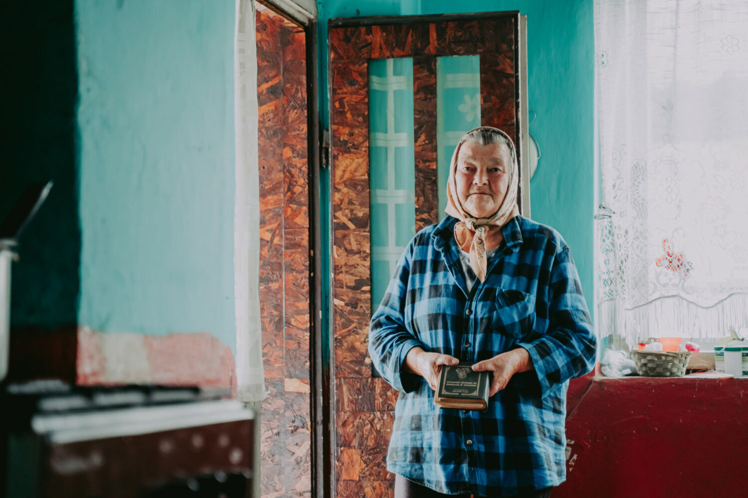 Een oudere vrouw staat binnen bij de deur, houdt een tablet vast en kijkt vriendelijk. Ze draagt een hoofddoek en een blauwe geruite blouse, in een gezellige, huiselijke omgeving.