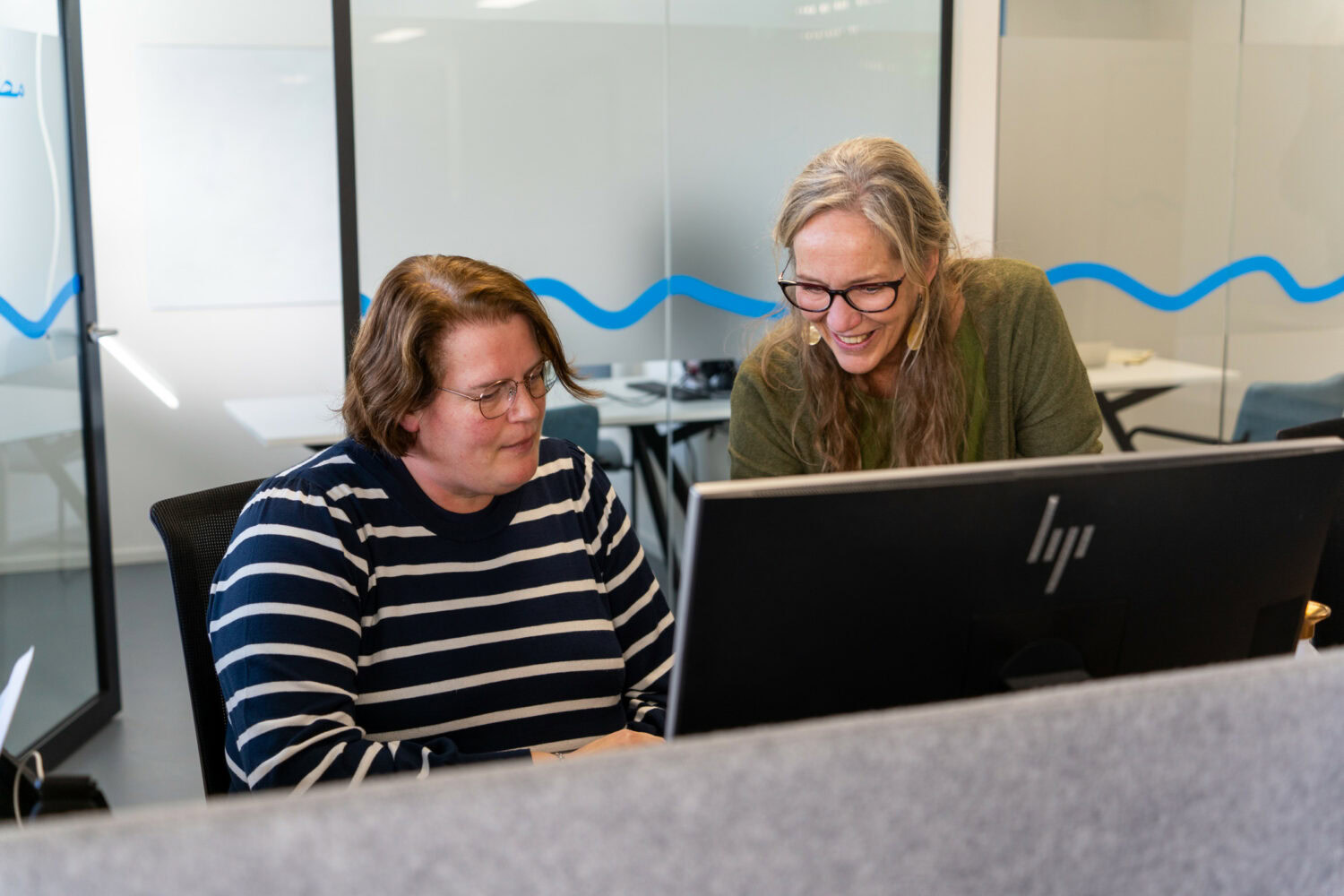 Twee vrouwen werken samen aan een computer in een kantoorruimte, lachen en bespreken iets, wat een professionele en vriendelijke werksfeer uitstraalt.