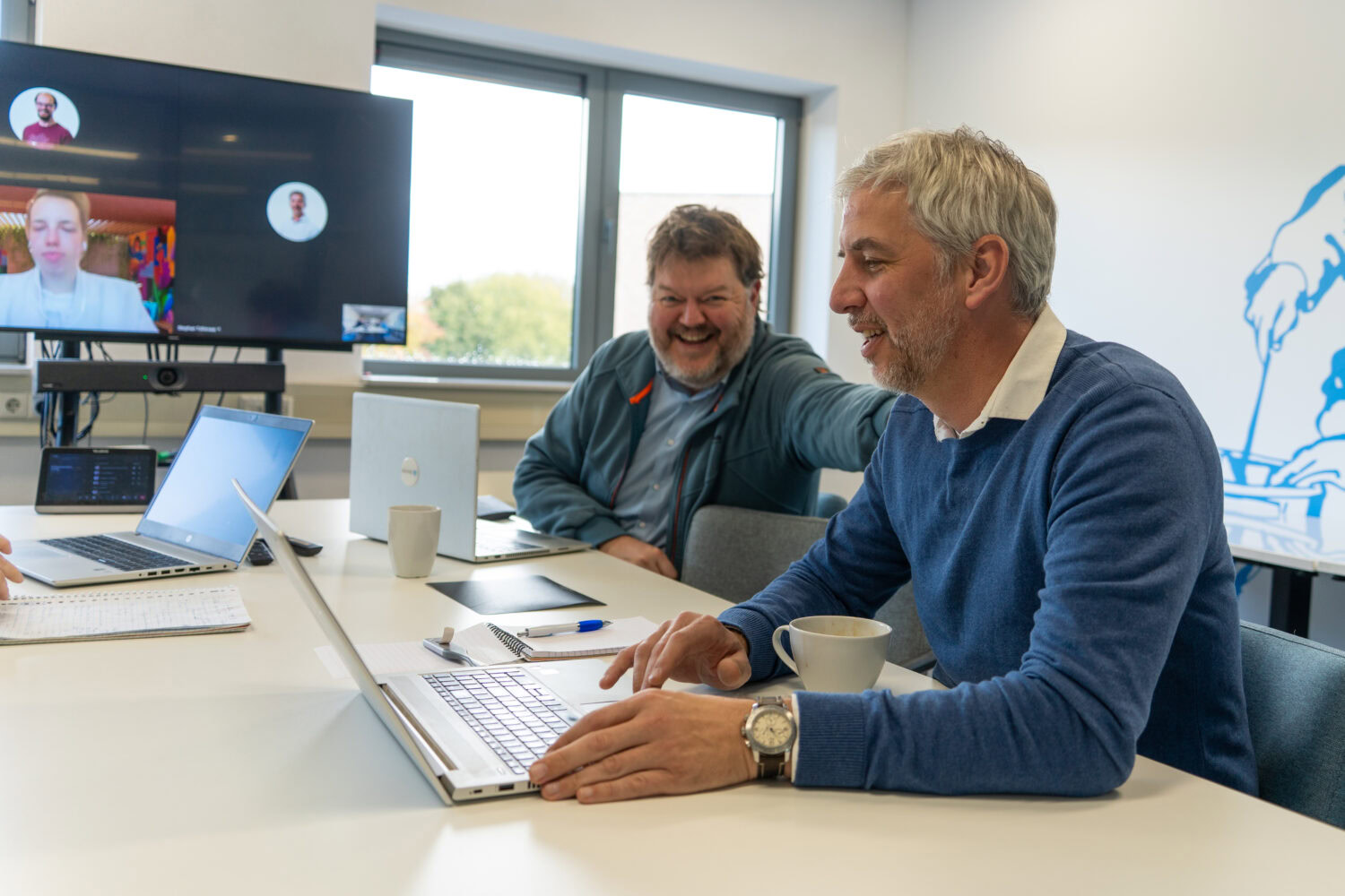 Een foto van twee mannen die samenwerken en lachen tijdens een vergadering op kantoor, met laptops en een scherm met videovergadering, onder het thema 'Kom in actie voor Dorcas'.