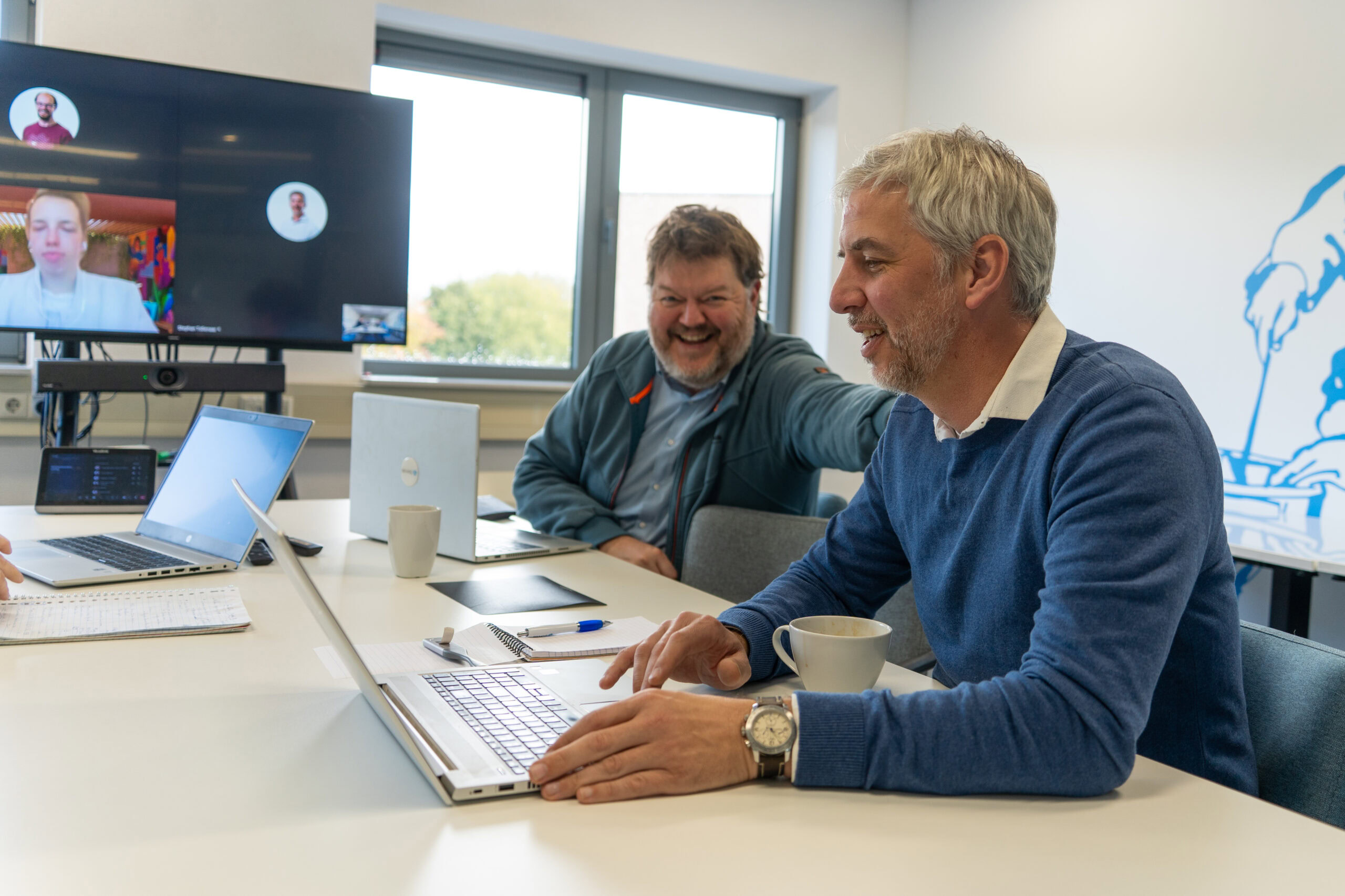 Een foto van twee mannen die samenwerken en lachen tijdens een vergadering op kantoor, met laptops en een scherm met videovergadering, onder het thema 'Kom in actie voor Dorcas'.