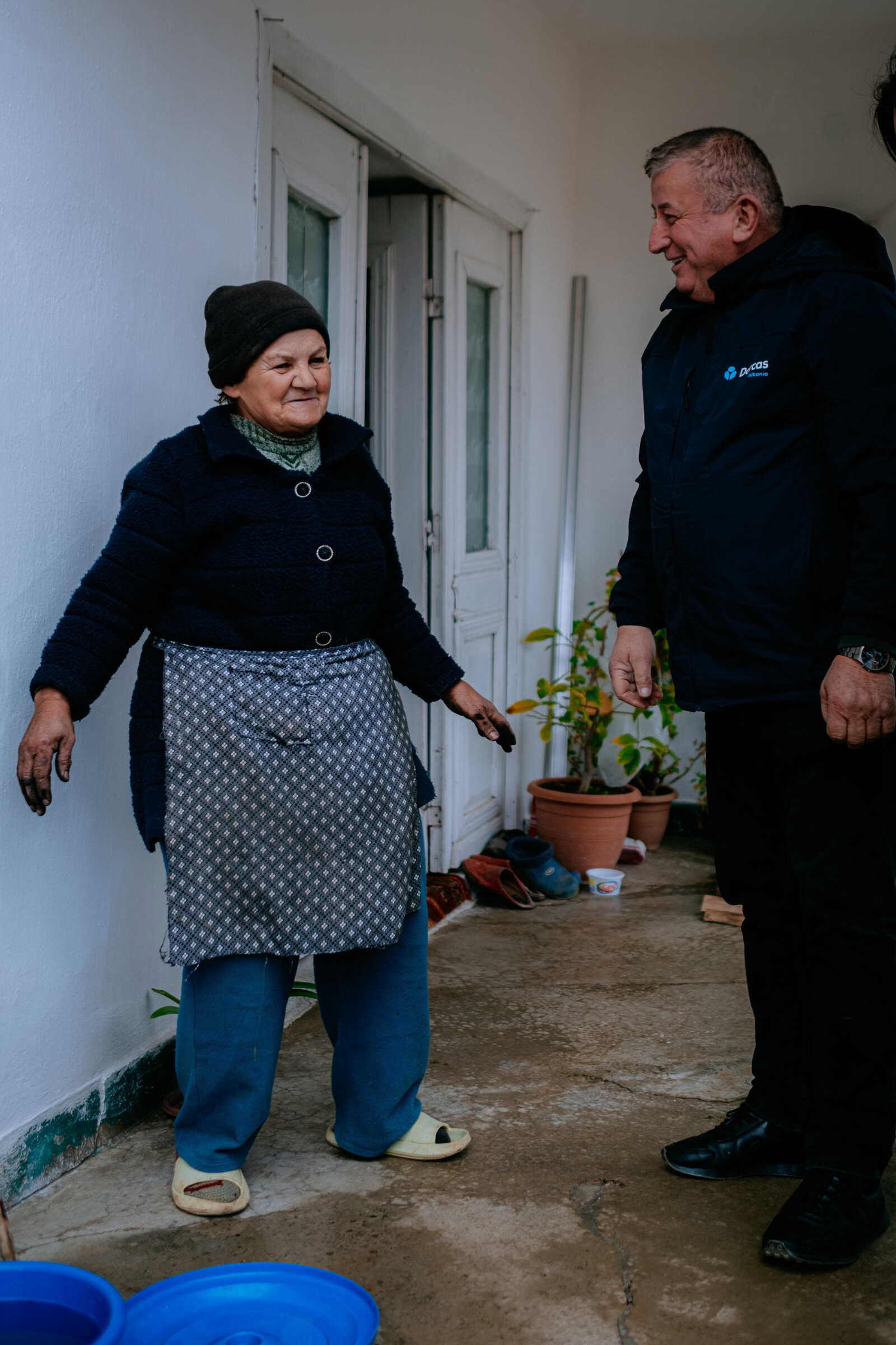 Hulpverlener spreekt met oudere vrouw in huis, beiden glimlachen, ondersteuning en zorg voor ouderen, betrokkenheid en warmte, binnenhuis, dorcas.nl.