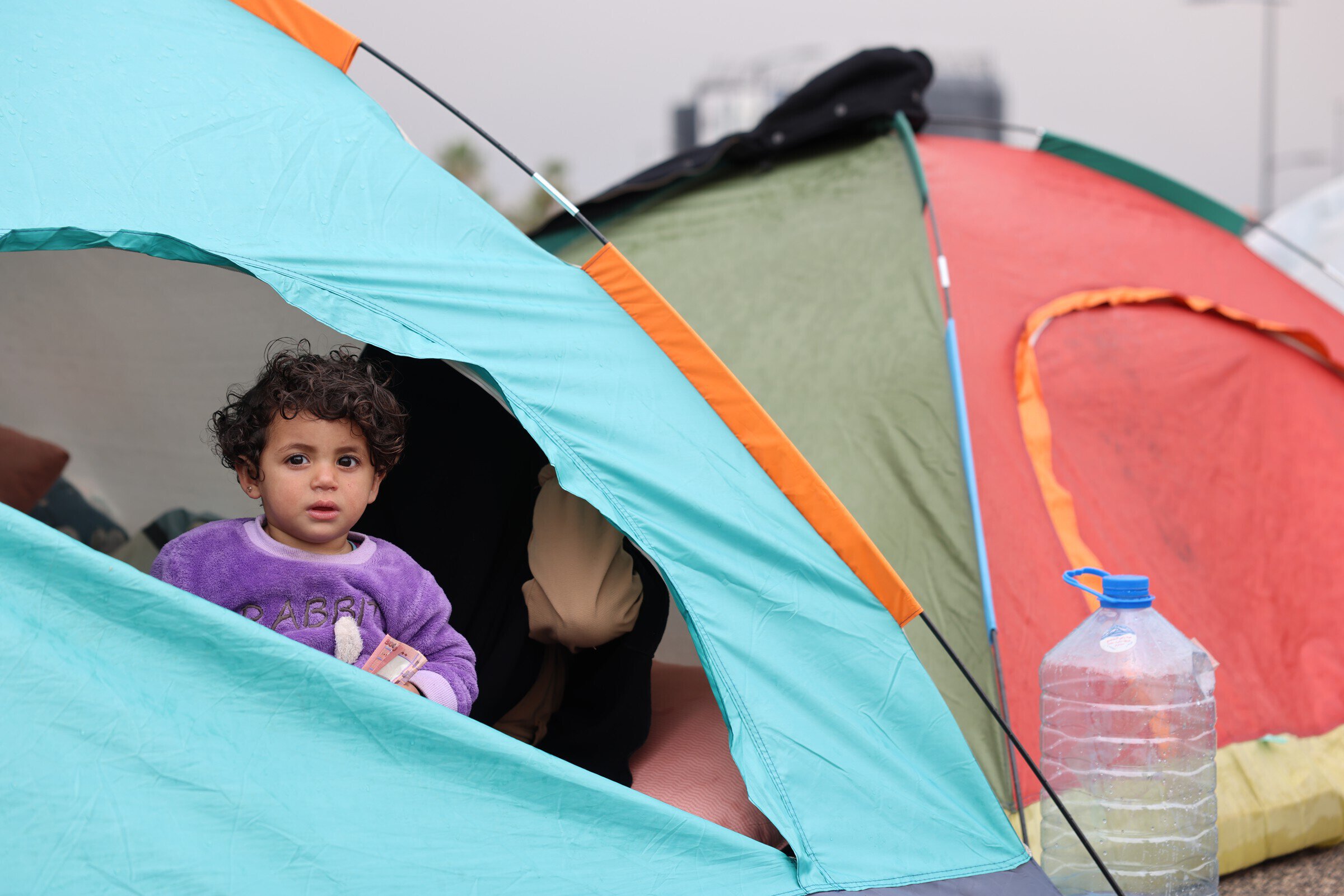 Een kind zit in een tent tijdens een noodhulpactie, met grote waterfles op de grond, symboliseert noodhulp en opvang voor getroffen mensen.