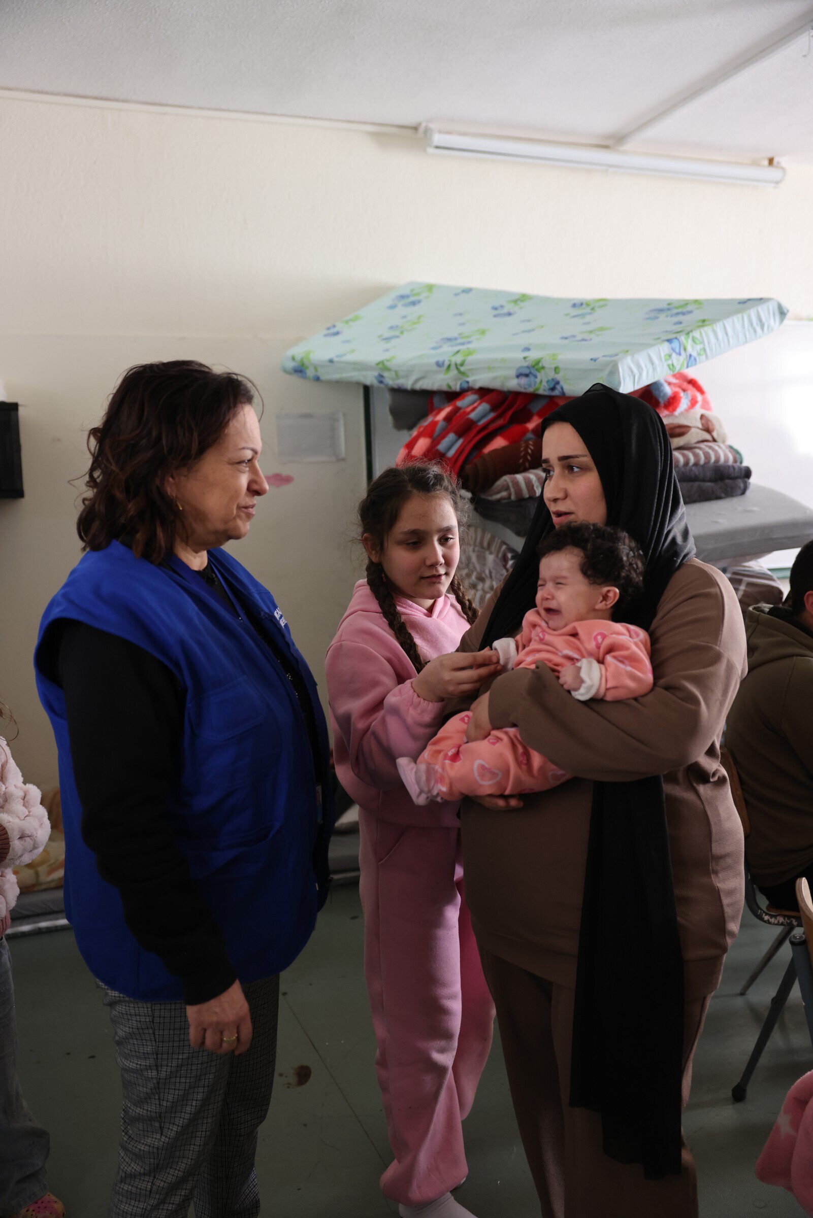Vrouw met hoofddoek houdt baby vast terwijl jonge meisje en hulpverlener in gesprek zijn, binnen een opvangcentrum voor vluchtelingen of migranten.