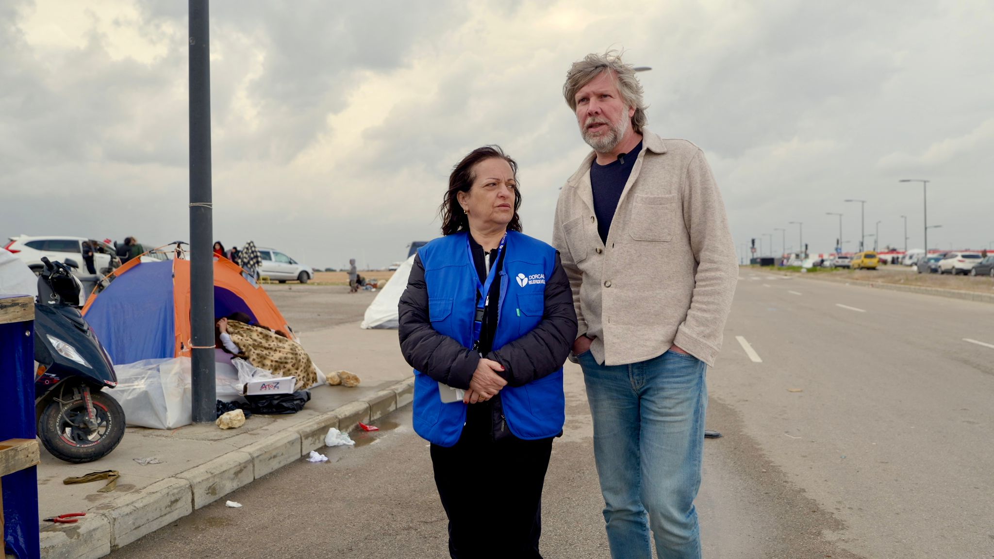 Een vrouw en een man staan samen op straat, omringd door tenten en persoonlijke bezittingen, terwijl ze betrokken en zorgzaam kijken.