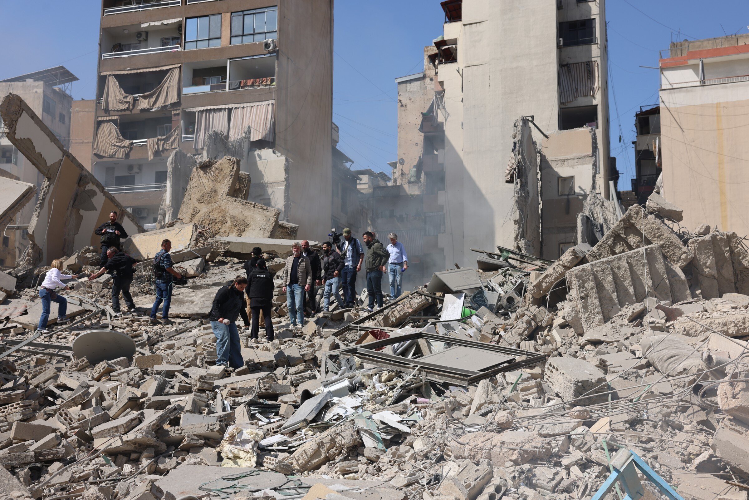 Mensen verzamelen zich bij de ruïnes van ingestorte gebouwen na de aardbeving in Beirut, met puin en stof op de achtergrond.