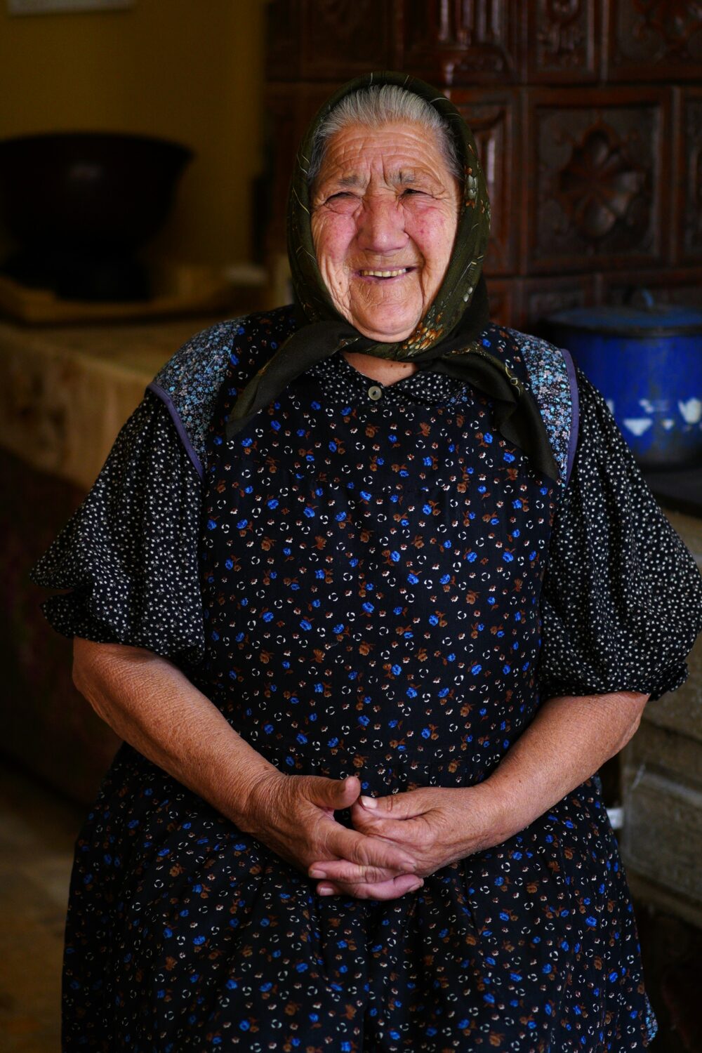 Oudere vrouw met hoofddoek glimlacht in haar huis, symboliseert warmte en zorg, passend bij het thema 'Sponsor het leven' en het ondersteunen van ouderen in de gemeenschap.