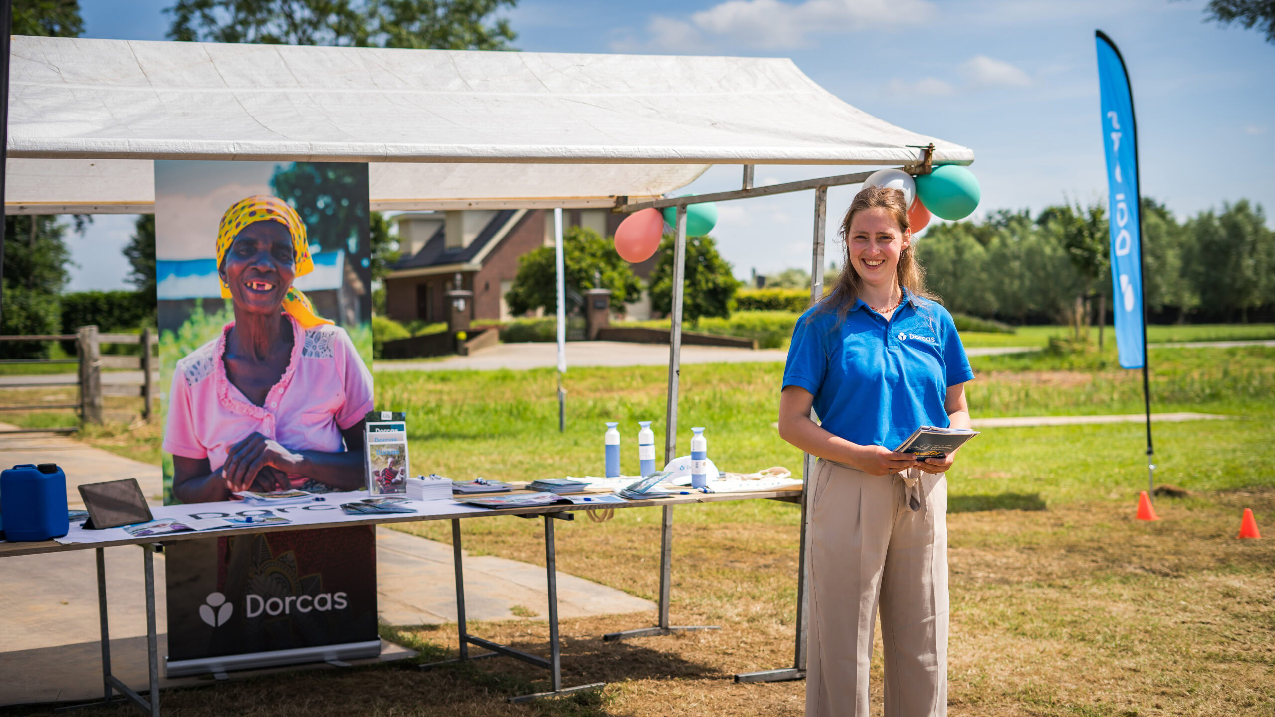 Een vrijwilliger staat bij een dorcas informatiestand onder een witte tent, met een lachende vrouw in een blauwe polo die een tablet vasthoudt, op een zonnige dag in een groene buitenomgeving.