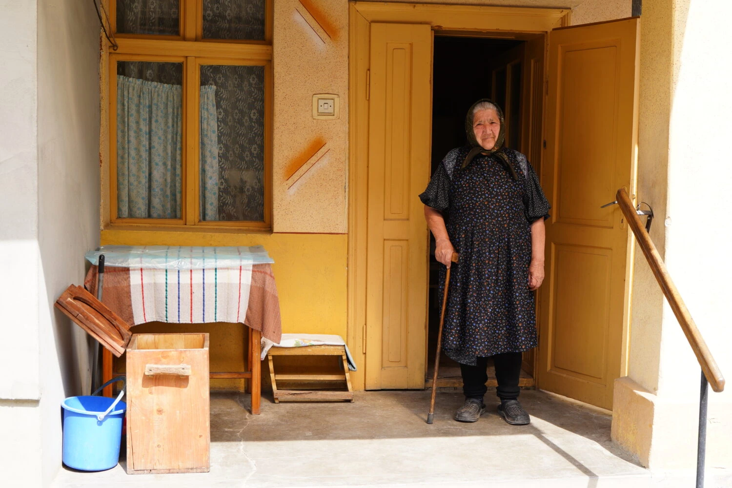 Een oudere vrouw staat met een wandelstok voor de open voordeur van haar huis, lachend en vriendelijk kijkend, in een gezellige en huiselijke omgeving.