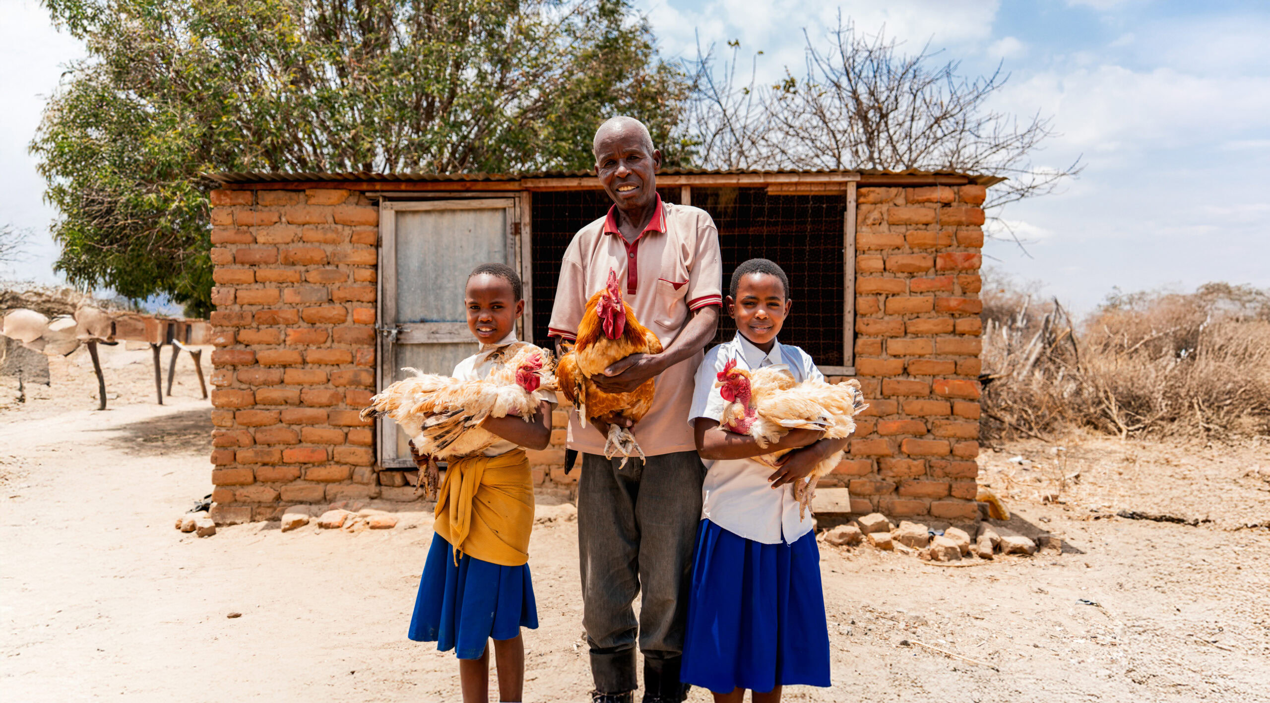 Een man en twee jonge meisjes met voedselpakketten voor kinderen in Kenia, in de buurt van een eenvoudige bakstenen woning, onder een heldere hemel.