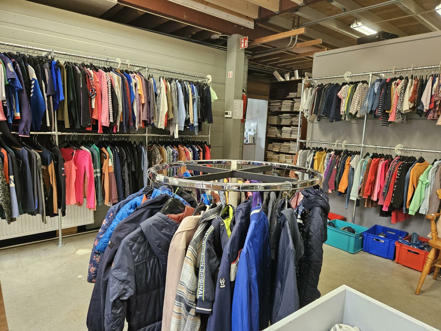 Kledingwinkel met diverse kledingstukken op hangers en rekken, georganiseerd in een ruime opslagruimte met houten plafond en plastic opbergboxen op de achtergrond.