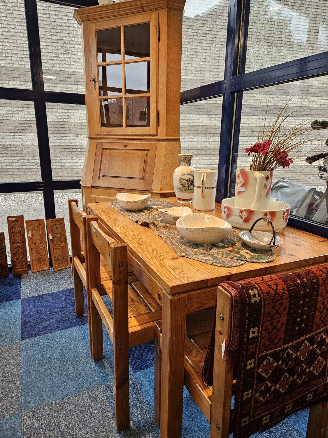 Houten eettafel met servies en decoratie, omgeven door houten stoelen, in een zonnige serre met grote ramen en een kleurrijk tapijt op de vloer.