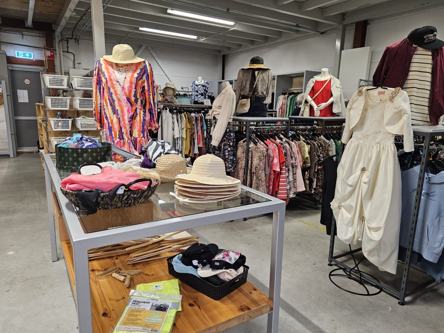 Zomerse kleding en accessoires uitgestald in een tweedehandswinkel, met hoeden, jurken en tassen op een winkelvloer.