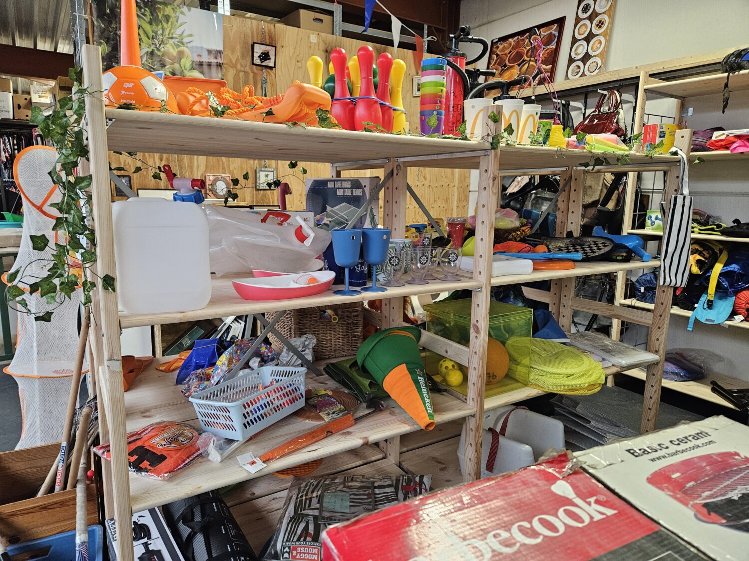 Zomerse strandspullen en speelgoed op een houten winkelrek, inclusief waterballonnen, emmers, schepnetten en zwembadspeelgoed, in een winkel met diverse andere artikelen op de achtergrond.
