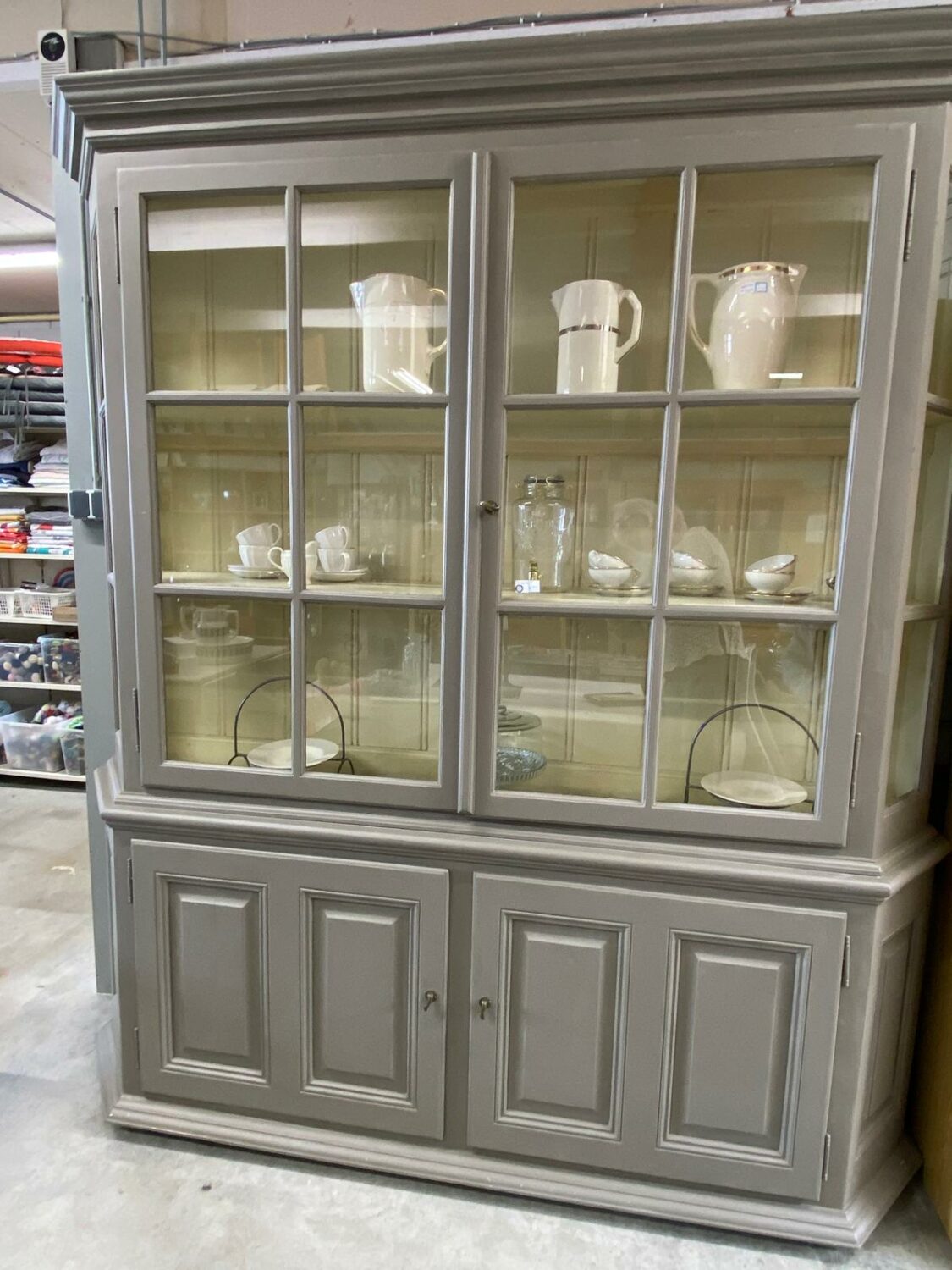 Zilveren houten vitrinekast met glazen deuren en meerdere witte serviesstukken, inclusief kommen, borden en kannen, in een magazijn of winkelomgeving.