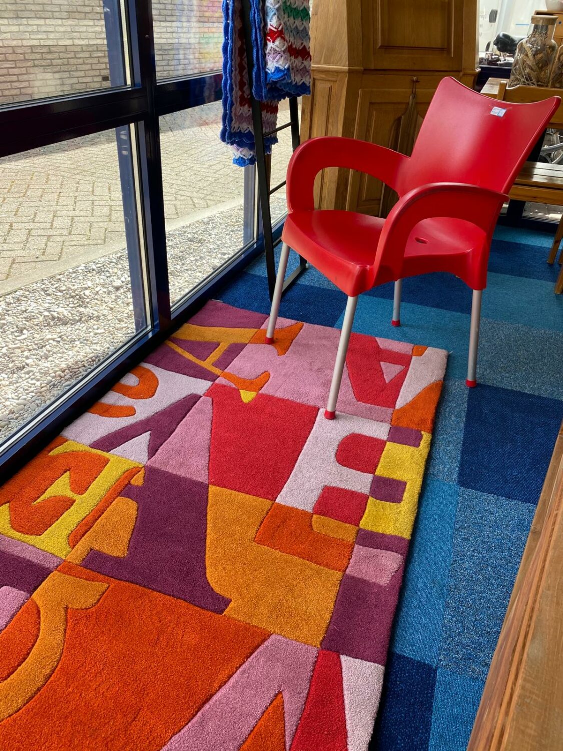 Een rode stoel naast een kleurrijk tapijt in een interieur met grote ramen en houten meubels.