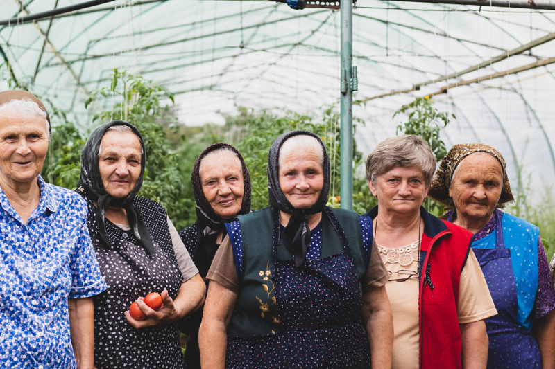 Vrouwen in een kas in Roemenië, gekleed in traditionele kleding, staan samen en glimlachen, met tomaten in de hand, tijdens een lokale landbouwactiviteit.