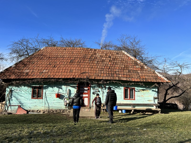Een groep mensen loopt naar een oud, traditioneel Roemeens huis met een rieten dak, gelegen in een landelijke omgeving onder een heldere blauwe hemel.