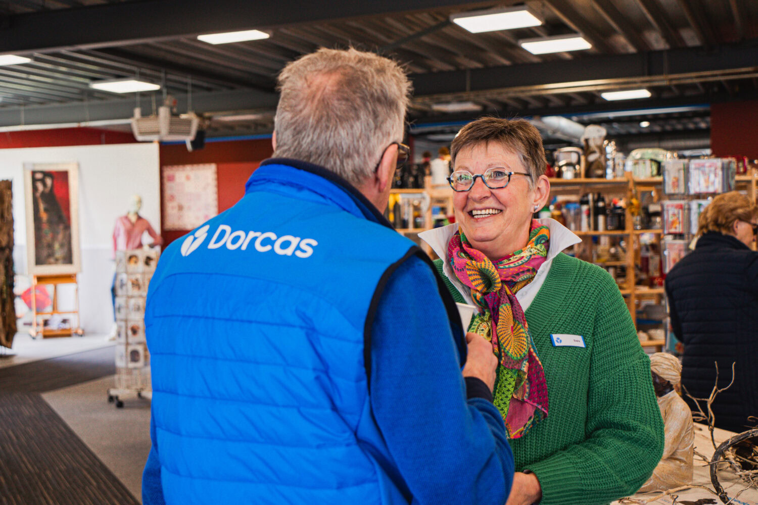 Een medewerker van Dorcas in een blauwe jas praat met een lachende klant in een kringloopwinkel, omringd door diverse tweedehands artikelen en een vriendelijke sfeer.