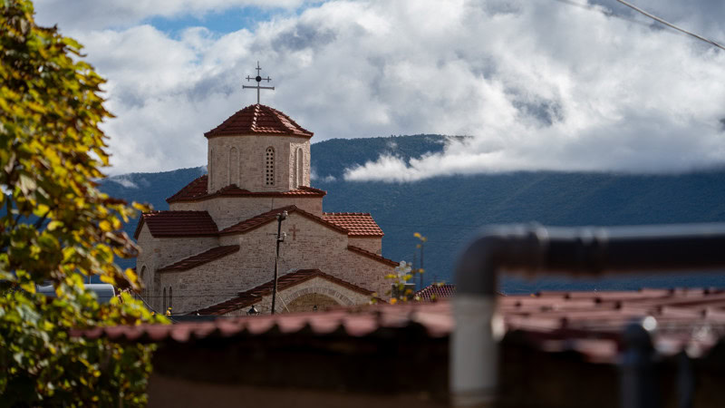 Albanië, klein kerkje met rode dakbedekking en bergachtig landschap op de achtergrond, onder een bewolkte hemel.
