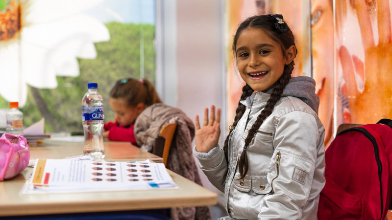 Kinderen in een klaslokaal met schoolspullen, waaronder een waterfles en een boek, terwijl een meisje vriendelijk naar de camera lacht en zwaait.