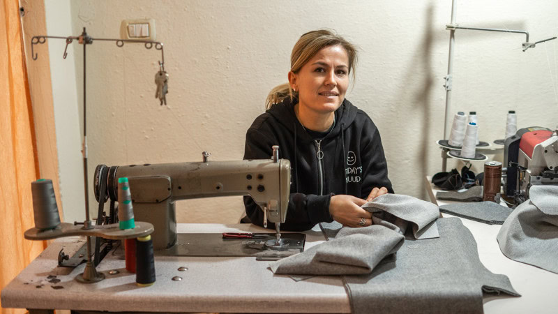 Een vrouw die in een naaiatelier werkt, omringd door stoffen en naaimachines, met een vriendelijke uitstraling.