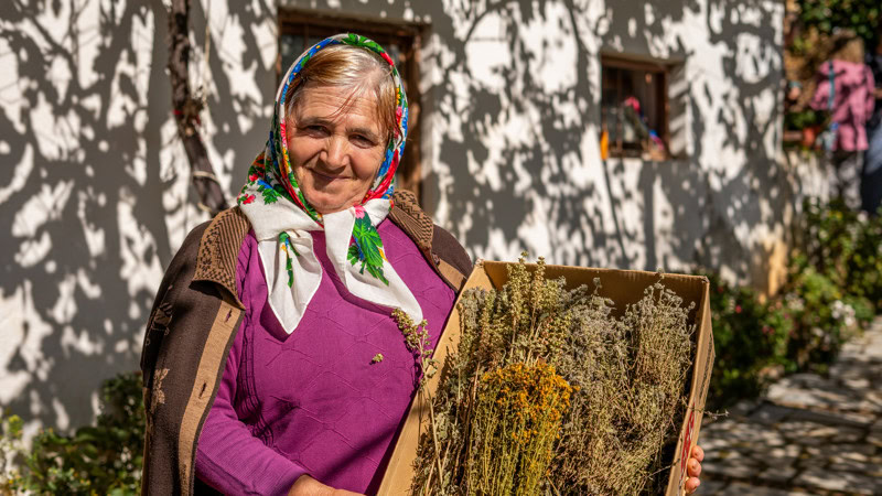 Een Albanese vrouw met een kleurrijk hoofddoekje en een mand vol bloemen, wandelend door een dorp met witte huizen en bloemen langs de straat.