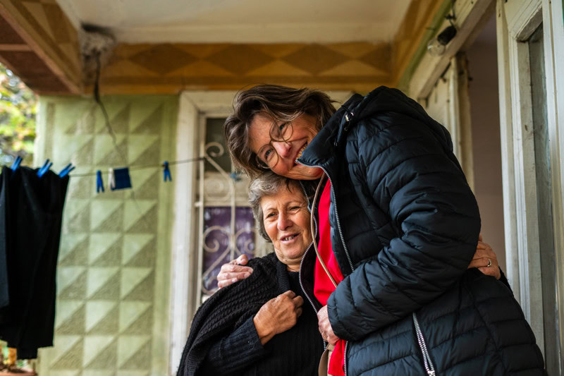Een warme omhelzing tussen twee vrouwen, lachend en knuffelend, in een huis met groene muur en waslijn op de achtergrond, uitdrukking van vriendschap en verbondenheid.