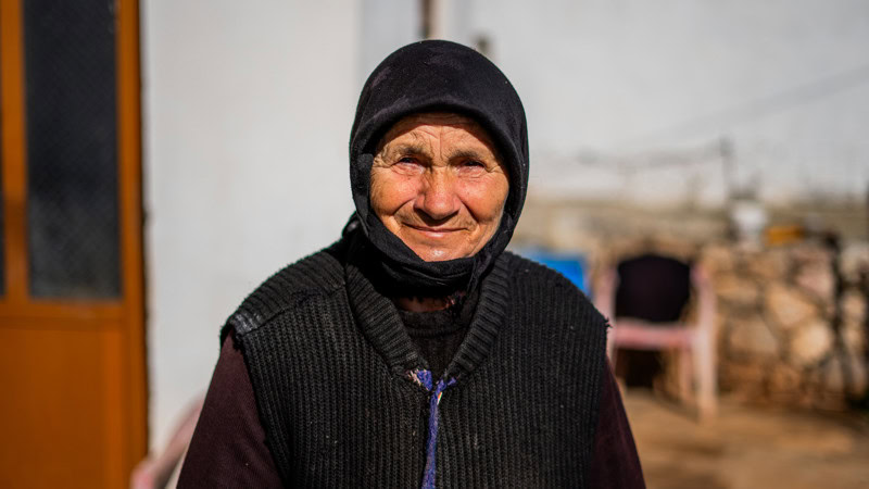 Een Albanese vrouw in traditionele kleding, lachend en warm gekleed, staat buiten in een landelijke setting met een bakstenen muur en houten deur op de achtergrond.