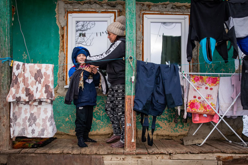 Een moeder geeft kleding aan haar kind voor de deur van een eenvoudige woning in Moldavië, waar armoede en gebrek aan basisvoorzieningen zichtbaar zijn.