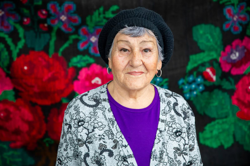 Een oudere vrouw uit Moldavië met een vriendelijke uitstraling, gekleed in een bloemenpatroon jas en een zwart hoedje, voor een levendige bloemenachtergrond.