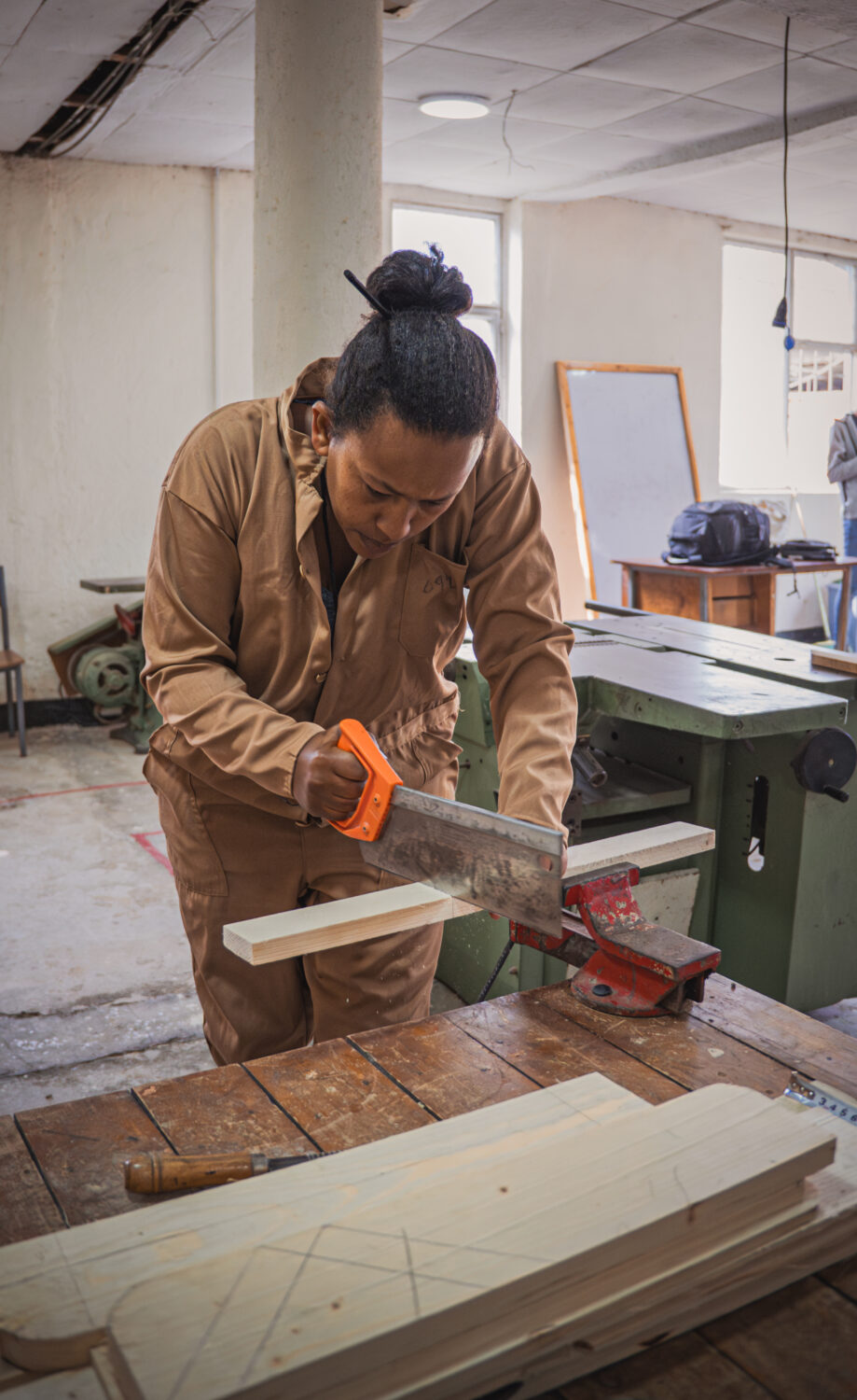 Een vrouw snijdt hout met een handzaag in een werkplaats, omgeven door hout en gereedschap, met een raam en werkbank op de achtergrond.